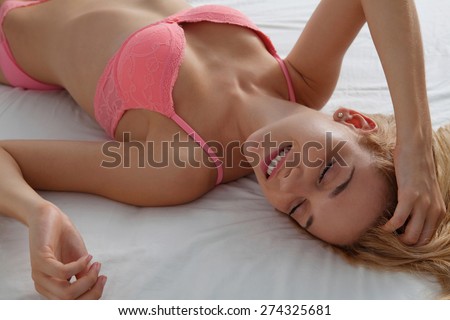 Overhead beauty portrait of young woman laying relaxing on a luxury bed in a home bedroom, smiling flirting wearing sexy pink bra lingerie indoors. Home lifestyle and skin hair care, interior.