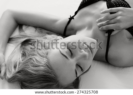 Black and white beauty portrait of young woman laying relaxing on a luxury bed in a home bedroom, smiling flirting, wearing sexy bra lingerie, indoors. Home lifestyle and skin hair care, interior.