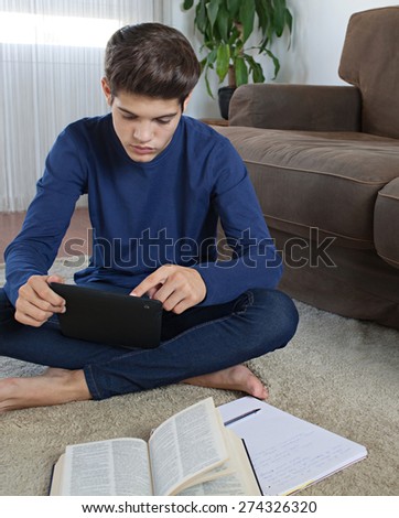 Teenager boy sitting on a rug in a living room using a digital tablet pad to do his homework and study at home. Young student man learning using telecommunications technology, home living lifestyle.