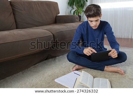 Teenager boy sitting on a rug in a living room using a digital tablet pad to do his homework and study at home. Young student man learning using telecommunications technology, home living lifestyle.