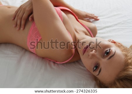 Overhead beauty portrait of young woman laying relaxing on a luxury bed in a home bedroom, smiling flirting, wearing sexy pink bra lingerie, indoors. Home lifestyle and skin hair care, interior.