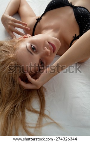 Over head close up beauty portrait of young woman laying on a luxury bed in a home bedroom, smiling flirting, wearing sexy black bra lingerie, indoors. Home lifestyle and skin hair care, interior.