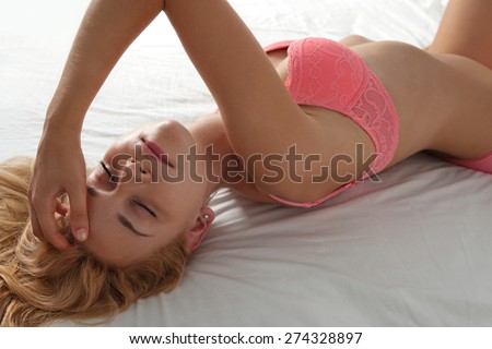 Beauty portrait of young woman laying relaxing on a bed in a home bedroom, flirting  with eyes shut, wearing sexy pink bra lingerie, hotel room, indoors. Home lifestyle and skin hair care, interior.
