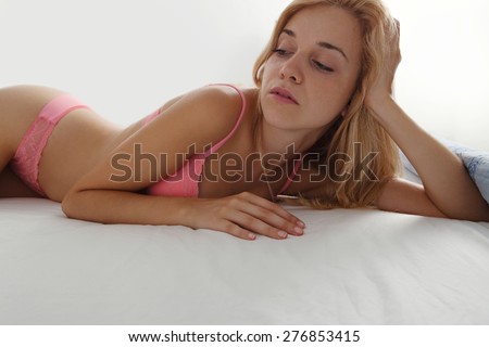 Beauty portrait of young woman laying relaxing on a bed in a home bedroom, thoughtfully looking away, wearing sexy pink bra lingerie, hotel room, indoors. Home living lifestyle skin care, interior.