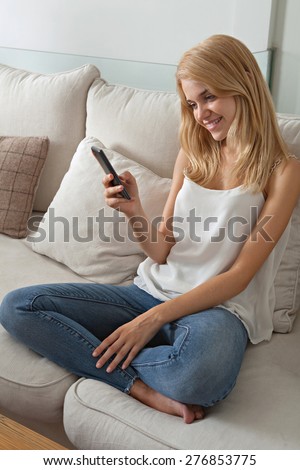 Attractive young woman sitting on a sofa at home, using a smartphone technology, networking on cell phone, smiling. Student girl using mobile phone internet at home, interior. Technology lifestyle.