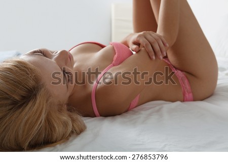 Beauty portrait of young woman laying relaxing on luxury bed in home bedroom, smiling while wearing sexy pink bra lingerie, hotel room, indoors. Home lifestyle and skin, hair and body care, interior.