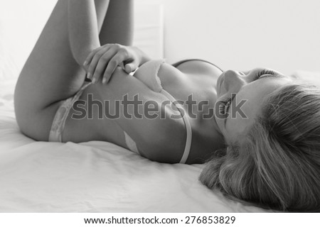 Black and white beauty portrait of young woman laying relaxing on bed in a home bedroom, wearing sexy bra lingerie, hotel room, indoors. Home lifestyle and skin, hair and body care, interior.