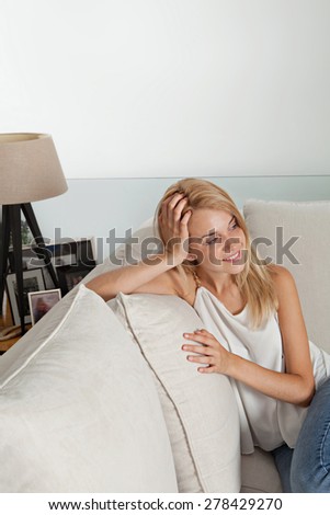 Portrait of a young beautiful teenager woman sitting and relaxing on a white sofa in a home living room, smiling and lounging at home. Luxury living lifestyle, interior. Young woman at home, happy.