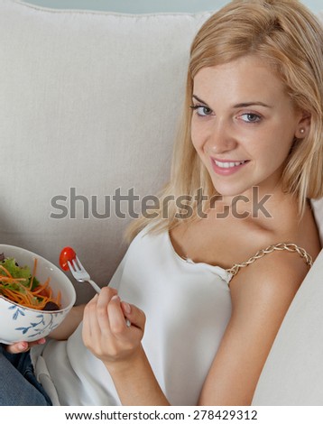 Portrait of attractive young woman sitting on sofa in a home family room living room, eating a fresh colorful salad, smiling. Girl eating healthy food at home, interior. Well being lifestyle.