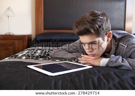 Young teenager man laying on bed at home, relaxing using a digital tablet to go on-line, interior. Lifestyle technology at home. Young person using telecommunications technology indoors, thoughtful.
