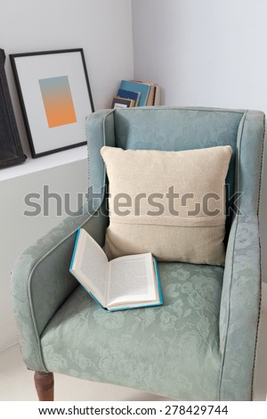 Still life home interior view of an elegant armchair in a stylish home with cushions and an open book laying on the chair, indoors. Reading room with picture frames and book, aspirational lifestyle.