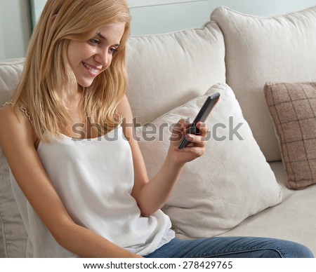 Attractive young woman sitting on a sofa at home, using a smartphone, networking on her cell phone, smiling. Student girl using mobile phone internet at home, interior. Home technology lifestyle.