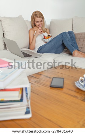Young woman relaxing on sofa in a stylish home living room, eating a fresh vegetable salad and using a laptop computer, indoors. Wellness, well being and healthy food. Technology lifestyle, interior.