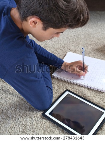 Rear side view of a teenager laying on a rug in a house living room using a digital tablet to study at home. Young student man learning using telecommunications technology, home living lifestyle.
