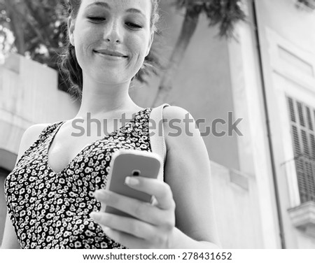 Black and white portrait of a beautiful young woman using a smart phone to go on line and network while on holiday, smiling outdoors. Girl using social media technology. Travel lifestyle exterior.
