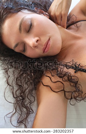 Portrait of beautiful young exotic woman laying on a bed at home relaxing and being sexy wearing sensual black lingerie with her eyes closed and a moody expression. Aspirational lifestyle, interior.