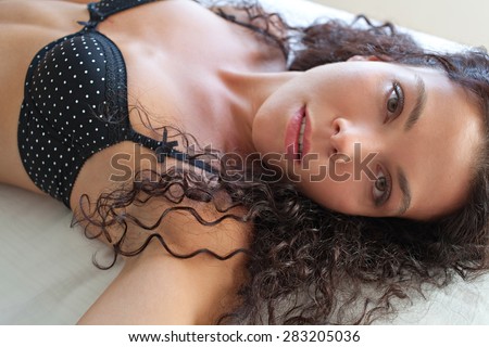 Portrait of a beautiful young exotic woman laying on a bed at home, being sexy wearing sensual black lingerie bra and looking at camera with a moody expression. Aspirational lifestyle, interior.