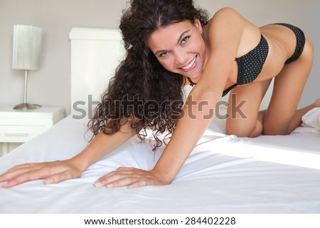 Beauty body figure of a young beautiful exotic sexy woman crawling on a bed at home in black lingerie, smiling, flirting and looking at camera in a bedroom, interior. Aspirational lifestyle, indoors.