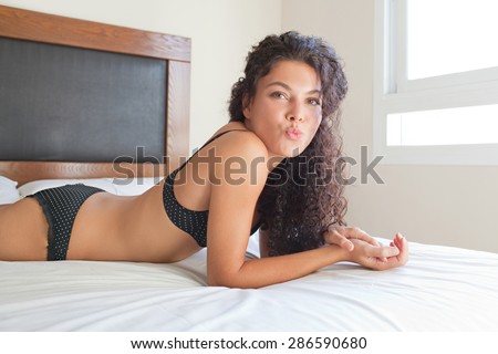 Side sexy body figure of a young exotic woman laying down on a bed at home in black lingerie, being sensual and playful and blowing a kiss to the camera, happy. Beauty in a home bedroom, interior.