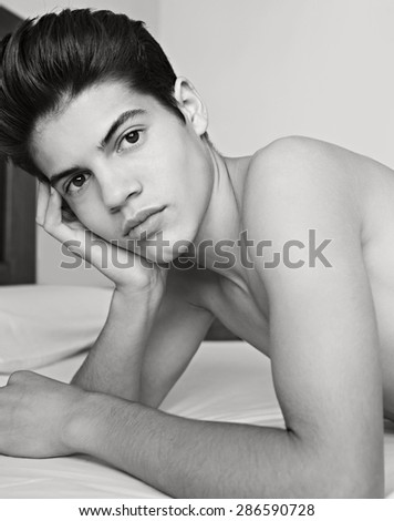 Black and white beauty portrait of a young adolescent teenager man laying on a bed at home with perfect skin, thoughtful and moody, indoors. Male puberty. Young person relaxing on a bed, interior.