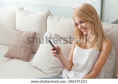 Portrait of beautiful young woman relaxing on a white sofa at home, using a smartphone to network on line, smiling in an elegant living room, interior. Aspirational lifestyle, indoors.
