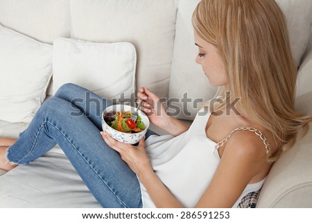 Beautiful young blond woman sitting on a sofa at home, eating a healthy salad in an elegant living room, home interior. Well being and aspirational lifestyle and healthy eating habits, indoors.
