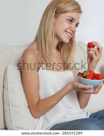 Beautiful young blond woman sitting on a sofa at home, smiling and eating healthy strawberries fruit food in living room, house interior. Well being lifestyle and healthy eating habits, indoors.