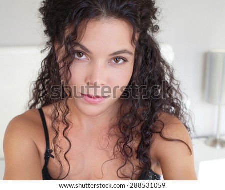 Close up beauty portrait and body figure of a young beautiful exotic sexy woman sitting on a bed at home, thoughtful and smiling, relaxing on a bedroom interior. Well being and woman issues, indoors.