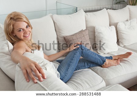Beautiful young blond woman sitting on a white sofa at home, smiling and lounging in a stylish and elegant living room, luxury spacious home interior. Aspirational lifestyle in quality house, indoors.