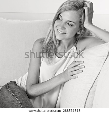 Black and white portrait of beautiful young woman relaxing on a white sofa at home, smiling in an elegant living room, luxury spacious home interior. Aspirational lifestyle in quality house, indoors.
