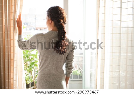 Rear view of a young woman wearing a robe and holding the curtains open to look out of a large light window at home, interior. Positive and aspirational lifestyle. Woman looking out a window, indoors.