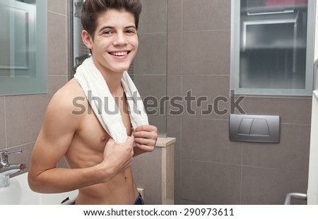 Beauty portrait of a young man in a home bathroom with a white towel around his neck, joyful and looking at camera, interior. Health, care and male grooming, indoors. Man portrait with perfect skin.
