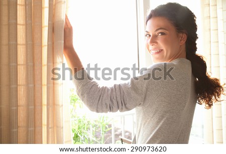 Rear view of a young joyful woman wearing robe and holding the curtains open to look out of large light window at home, turning to look and smile at camera, interior. Aspirational lifestyle indoors.