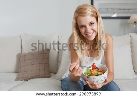 Beautiful young blond woman sitting on a white sofa at home, smiling and eating a healthy salad in an elegant living room, home interior. Well being lifestyle and healthy eating habits, indoors.