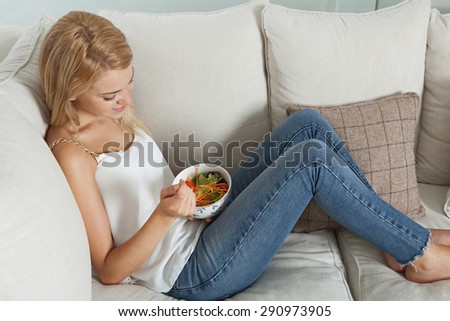 Profile view of a young woman sitting on a white sofa at home, eating a healthy salad food in an elegant living room, relaxing in interior. Well being lifestyle and healthy eating habits, indoors.
