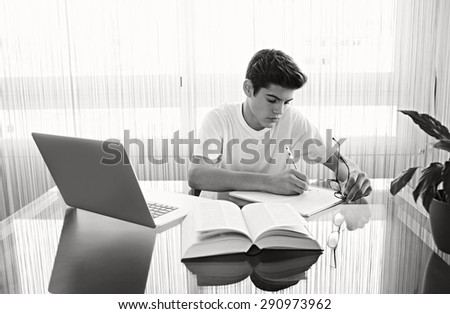 Adolescent teenager man doing his college home work at home, using a laptop computer, sitting at a glass reflective desk with large window, home interior. Technology student lifestyle indoors.