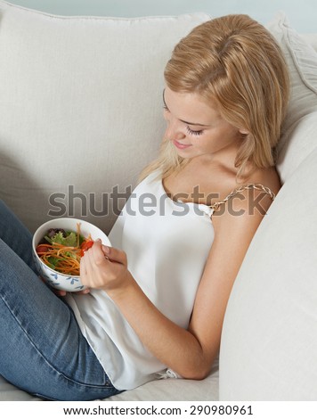 Beautiful young woman sitting on a white sofa at home, eating a healthy salad food in an elegant living room, relaxing in luxury interior. Well being lifestyle and healthy eating habits, indoors.