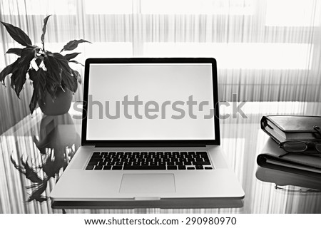 Black and white still life of office room with open laptop computer on glass desk with reflections by large bright window, office interior. Professional home workplace, technology indoors.