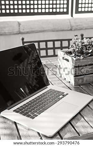 Black and white still life of open laptop computer on a terrace wooden table with flower pot in home exterior area, outdoors. Technology at home and working from home environment with no people.
