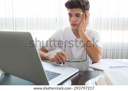 Teenager boy at a glass desk at home doing his homework using a laptop computer, holding reading glasses being thoughtful, interior. Adolescent  using technology, studying by large window, indoors.