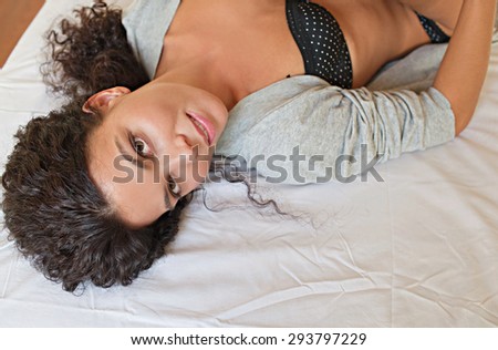 Over head beauty portrait of young attractive woman laying down on a bed at home, looking at the camera wearing lingerie and a robe, bedroom interior. Wellness, well being and healthy living indoors.