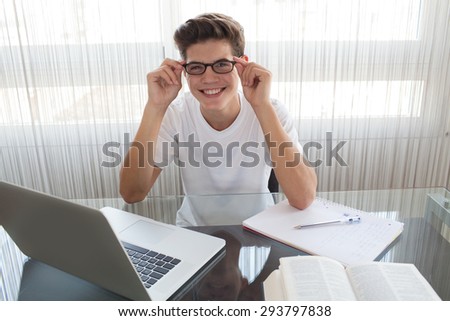 Adolescent young man at a glass desk at home using a laptop computer, wearing and holding his spectacles and joyfully smiling, interior. Teenager boy studying against large window, indoors.