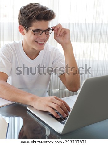 Adolescent young man at a glass desk at home doing his homework using a laptop computer, wearing spectacles and joyfully smiling, interior. Teenager boy studying against large white window, indoors.