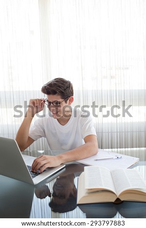 Adolescent young man at a glass desk at home doing his homework using a laptop computer, wearing spectacles and smiling preparing exams, interior. Teenager boy studying by large window, indoors.