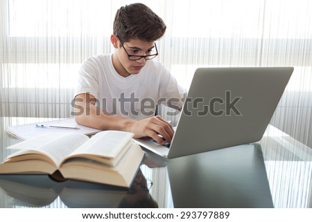 Adolescent teenager man at home, wearing reading glasses using a laptop computer, sitting at glass reflective desk with large window, home living room interior. Technology student lifestyle indoors.