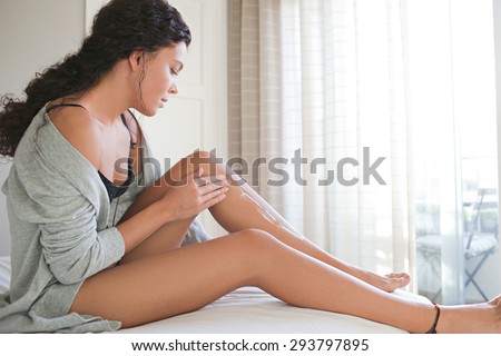 Side body view of a beautiful young woman applying lotion with her hands on her legs skin, smiling sitting on a white bed at home, in robe and lingerie, bedroom interior. Health wellness, interior.