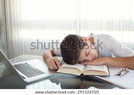 Teenager boy falling asleep at desk at home doing his homework using a laptop computer and books, being tired, interior. Adolescent using technology, studying by bright window with curtains, indoors.