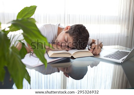 Teenager boy sleeping at a desk at home doing his homework using a laptop computer and books, being tired, interior. Adolescent using technology, studying by bright window with curtains, indoors.