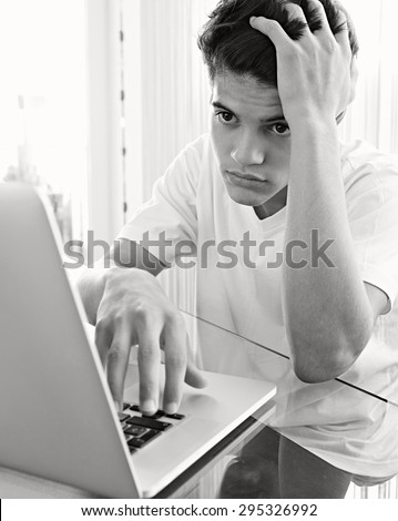 Black and white portrait of teenager boy at desk at home doing his homework using a laptop computer, typing and being doubtful, interior. Adolescent using technology, studying by window, indoors.