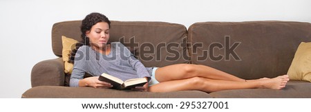 Beautiful young woman lounging on a comfortable sofa at home, reading a book while on holiday, living room interior. Relaxing home living lifestyle, indoors. Girl reading a book on the weekend.